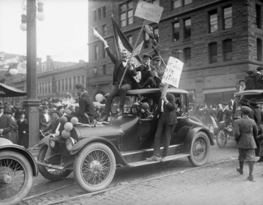 World War One military recruits ride a car in a Denver, Colorado, parade. The men hold signs, United States flags, blow party horns and wave to the crowd. Picket sign reads: "The Kaiser Has;" a dead duck hangs from it. Office buildings are in the background.