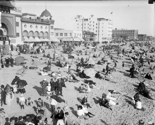 People with umbrellas crowd Santa Monica Beach in Santa Monica (Los Angeles County) California. Shows businesses and the Ocean Park Bath House, a Moorish style building with arches and domes.