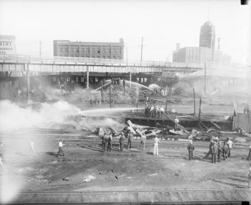 Fire fighters battle a blaze at the McPhee - McGinnity Lumber Company lumber yard under the Broadway viaduct in Denver, Colorado, as people look on. The McPhee-McGinnity building with clock tower, and "Pittsburgh Plate Glass" are in the background.