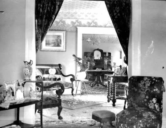 Interior view of a Denver, Colorado residence; decor includes brocade upholstery, a settee, chairs, tables, a foot stool, china pitchers, crystal, carpet, rugs, and a potted palm.