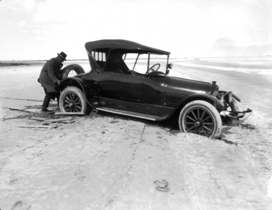 View of a car mired in mud by a river, (probably) in Colorado; a man lifts the spare tire from the back of the vehicle.