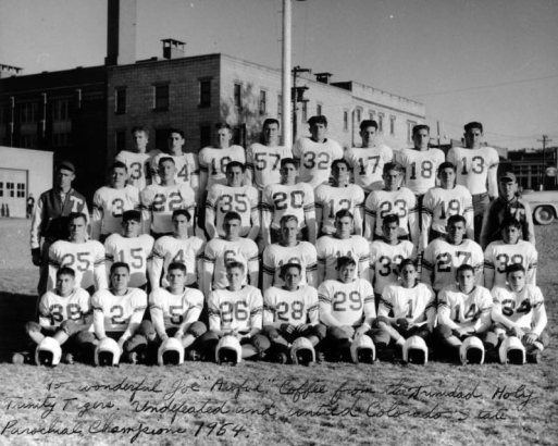 Portrait of a football team in Trinidad, Colorado. Teenage boys and young men in white, numbered jerseys are posed, the ones sitting in the front have their helmets on the grass in front of their cleated shoes. Two men in ballcaps and jackets with large "T" patches flank the standing row. Power poles and a three-story, flat roofed cream colored brick building is in the background.