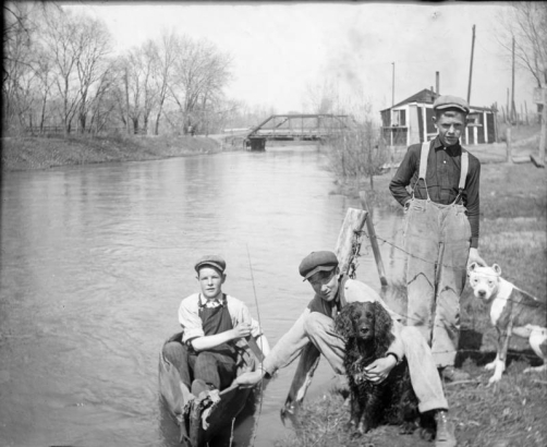 Outdoor portrait of boys with their dogs by Archer Canal in Denver, Colorado. One boy is in a canoe. Charles S. Lillybridge's house and studio and the Alameda Avenue bridge are in the distance.