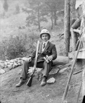 Photographer Harry Rhoads sits on a rock in a suit and pith helmet; he holds binoculars and a rifle; a pistol is in his jacket pocket, probably Colorado.