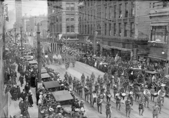 View of a parade in Denver, Colorado; shows marching soldiers and a band, cars, crowds, and United States flags.