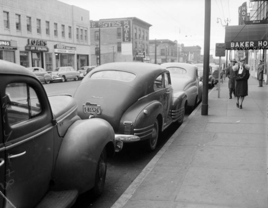 View of Welton Street in Denver, Colorado; shows parked cars, pedestrians, and business signs: "Jim Furlong Loans," "Baker Hotel," and "Estes Hotel."