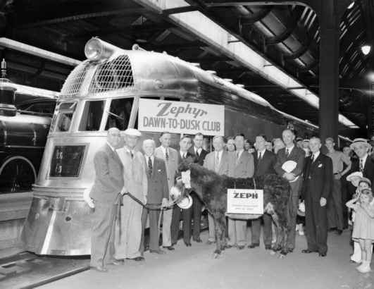 Men, boys, and girls pose with a burro in Chicago (Cook County), Illinois; and signs: "Zephyr Dawn to Dusk Club" and "ZEPH Denver to Chicago May 26, 1934." A Chicago, Burlington, and Quincy Railroad locomotive is in the background. Fred G. Gurley, later president of the Atchison, Topeka and Santa Fe, is fourth from the right, with the wire rim glasses.