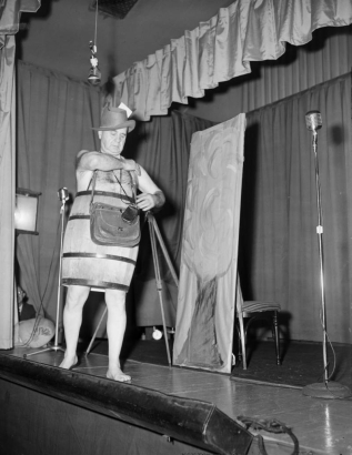 Portrait of photographer Harry M. Rhoads, on stage in (probably) Denver, Colorado, wearing a wooden barrel, a Fedora hat, and camera equipment bags. A microphone is to the side.