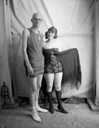 A man and a teenage girl pose in swimming suits; hers has a peacock motif, and she holds a shawl with fringe.