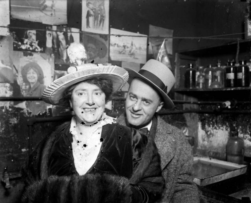 A man and woman pose. The woman wears a fur muff and fancy hat. Darkroom equipment and photographs are on interior walls.