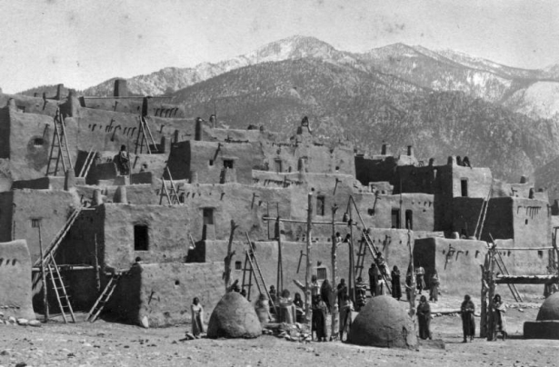 View of Taos Pueblo, New Mexico; shows adobe pueblo dwellings, hornos (bee-hive ovens), Pueblo Peak, and Native Americans (Taos).