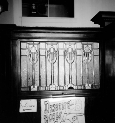 Interior view of a leaded glass window in the Columbian Hotel, in Trinidad, Colorado. Vertical panels of "ice" frosted glass contain leaf shapes interlocked with stirrup shapes at the top; a border with arches at regular intervals crosses the bottom. Signs under the window read: "Women" and "Dining Room".  Signs are illuminated by a shaded gallery lamp mounted on the polished wood frame that holds the window.
