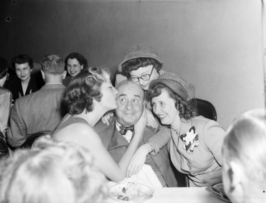 At a party, Frances Melrose (with corsage) laughs while Harry M. Rhoads is kissed on the cheek by other women.  Elizabeth "Liz" Wyner with glasses is behind Harry.  Denver, Colorado.