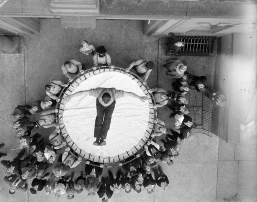 A man falls into a round Browder safety net at a fire department demonstration in Denver, Colorado. Men and Boy Scouts look on.