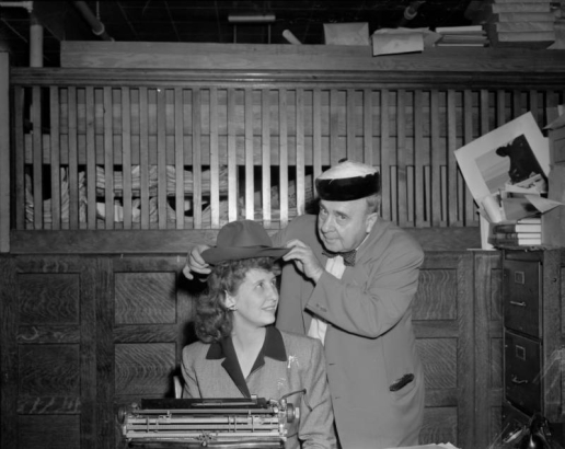 Harry M. Rhoads places a fedora hat on an Frances Melrose who is seated at a typewriter. Harry wears a chinese style hat, possibly a women's hat.