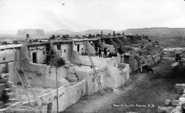 View looking east over Acoma Pueblo, Acoma Mesa, New Mexico, shows Pueblo Native Americans, multi-story adobe house blocks with ladders, vigas, chimneys, pottery, chickens, firewood and surrounding buttes and mesas.