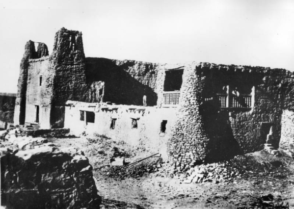View of the old mission church and convent at the Native American, Acoma Pueblo, show a few men posing on balconies of the weathered adobe structure.