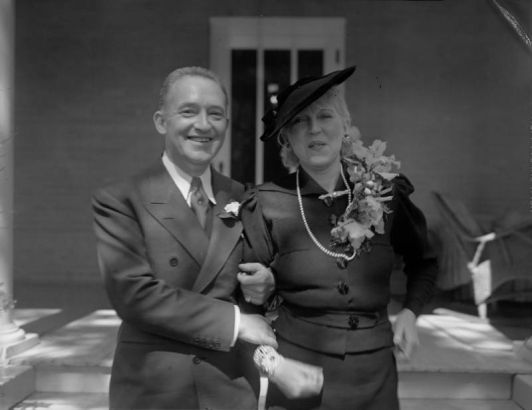 Newlyweds Helen Bonfils and George Somnes pose outside of a home. Helen wears a corsage, hat and string of pearls. The pearls may have been a gift given to her by Mrs. Harry H. Tammen, the hostess of the newlyweds wedding breakfast. George Somnes wears a suit, tie and a flower pinned on his lapel. Helen and George were married on September 15, 1936.