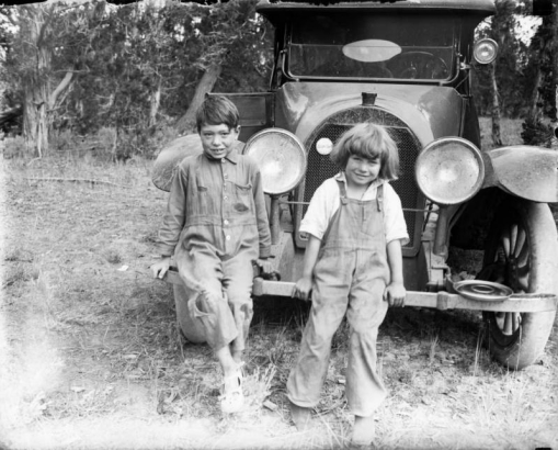 A boy and girl smile, sitting on an automobile bumper; she wears bib overalls.