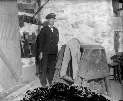 A Colorado State Penitentiary guard in uniform looks on as a man bends over the "Old Gray Mare," a device used in Canon City, Fremont County, Colorado, at the prison, for disciplinary beatings. Rolled United States flags are on an interior shelf.