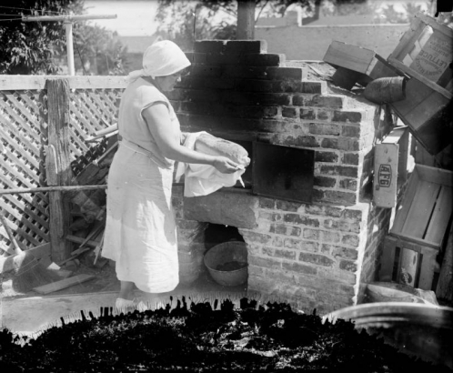 A woman holds a loaf of bread in a towel, by an outdoor brick oven.