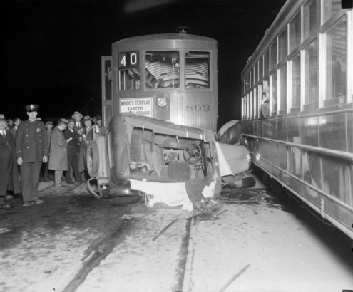 View of a car / streetcar crash; people look on, and signs read: "Knights Templar Easter Service," "40," and "803."