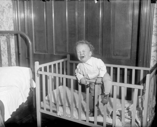 A small boy cries, climbing the side of his crib.