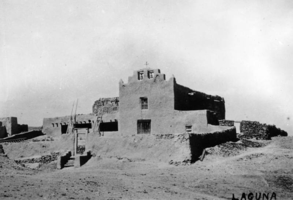 View of the Native American Laguna Pueblo Indian mission Church of San Jose, New Mexico, shows an adobe building with walled burial ground, stepped parapet, two bells and a cross.