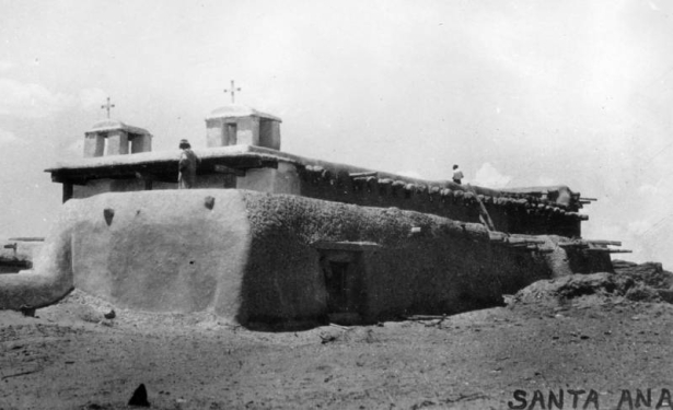 View of the mission church, Santa Ana Pueblo, New Mexico, an adobe building with vigas and twin belltowers topped with crosses.  Shows a Native American and a white man wearing a short sleeved shirt and hat.