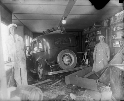 A man and woman pose by an automobile, wreckage, and interior store walls.