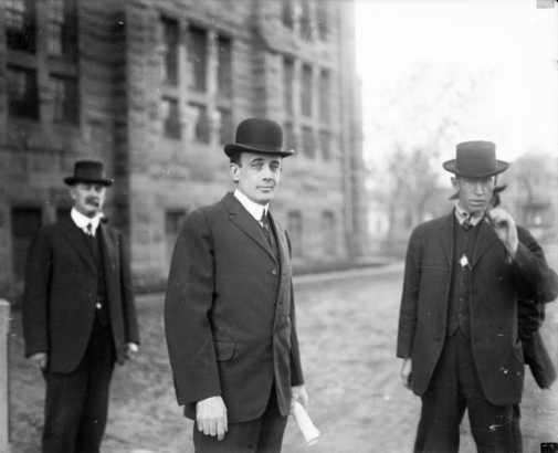 Three men stand outside of the Denver City Jail during the Brown Palace murder trial in Denver, Colorado. They wear suits, vests and hats. One man holds a cigar. The man in the center, with a rolled piece of paper in his hand, is the defendant, Frank Henwood.