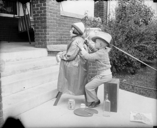 A girl sits on a stool in front of the porch of a home in Denver, Colorado. A boy is seated behind her and uses a paint brush on the back of the young girl's coat. On the ground is a palette with brushes, paint cans, and a bottle.
