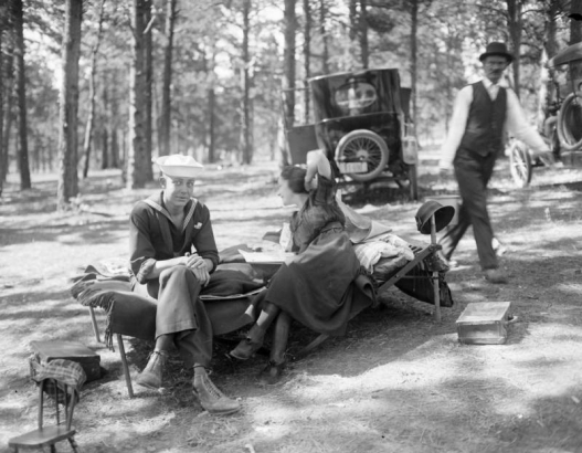 A man dressed in a sailor uniform and a woman enjoy a day outdoors as they sit on a cot in a park or forest, probably in Colorado. An automobile is parked in the background. A man walks by behind them.