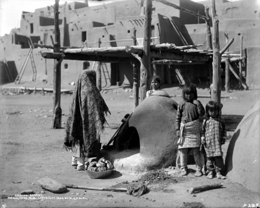 Native American (Pueblo) woman and three children stand by a beehive oven (horno), north plaza, Taos Pueblo, New Mexico. The woman wears a shawl covering her head and shoulders, there is a basket of bread by her feet. An adobe pueblo cluster is in the background.