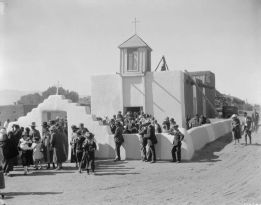 A crowd of men, women, and children exit an adobe church in Taos Pueblo (Native American), New Mexico. The church has a walled courtyard, stepped arch gate with a cross, wood steeple with a cross, and a bell on the roof. A car is parked in back.