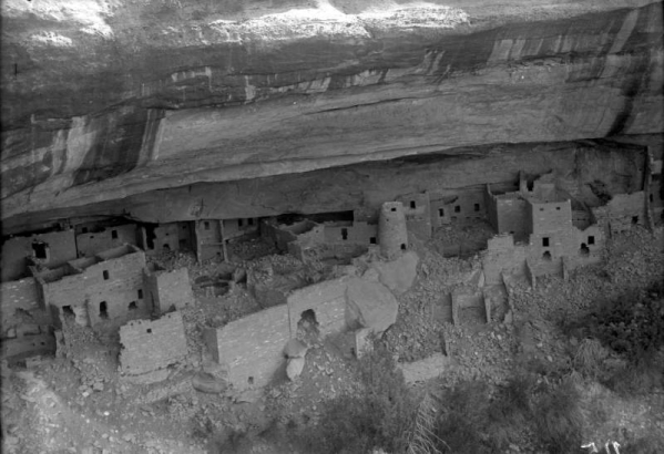View of Cliff Palace, a group of Native American (Anasazi) cliff dwellings, before excavation, Chapin Mesa, Mesa Verde National Park, Montezuma County, Colorado.  Window holes are visible throughout the masonry homes' walls. One wall has a large hole and shows signs of cracking. Masonry and rocks litter the grounds.