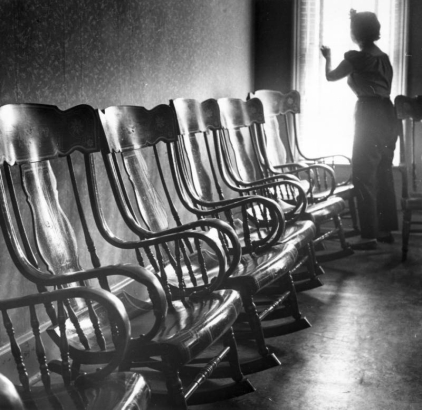 An interior view of the Clear Creek County Courthouse (?) in Georgetown, Colorado, including five Victorian rocking chairs lined up, a woman in slacks and a peasant blouse looking out the window, another rocking chair (partially visible), and wallpaper with small print. The chairs and floor shine from the window's light.