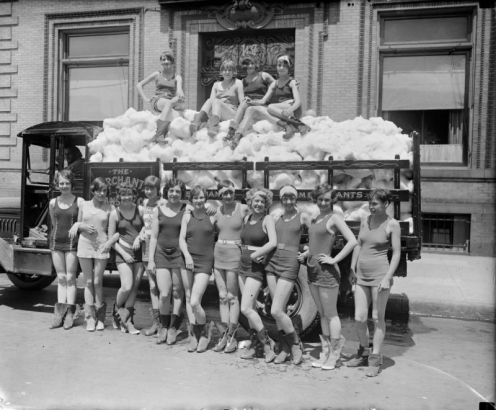 Young women and teenaged girls smile and pose in swimming suits in front of a panel truck full of snow or other material. Four of the women sit on the snow? or other material in the truck bed.