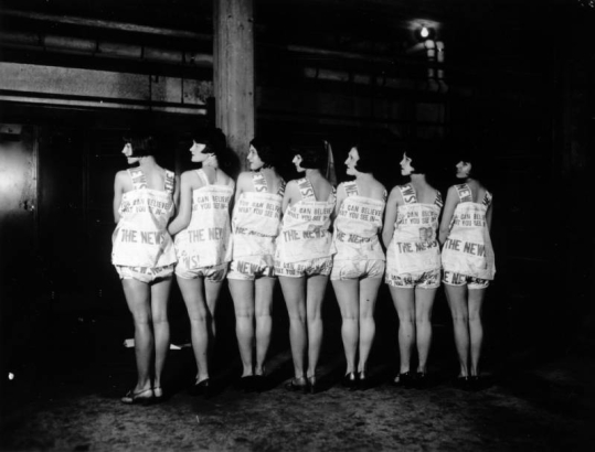 A dance team of women wear identical outfits printed with Rocky Mountain News pages; headline reads: "You Can Believe What You See In The News!"