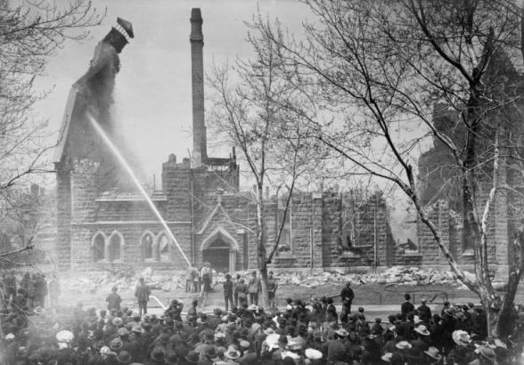 Firemen aim a jet of water at a collapsing chimney amid the ruins of the 23rd Avenue Presbyterian Church after the fire that destroyed the 1892 building in 1906. A crowd of spectators look at the remains of the stone building.