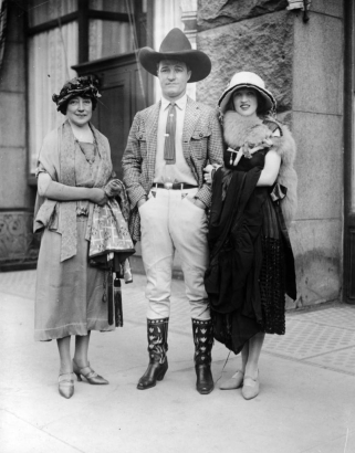 Actor Tom Mix poses with two unidentified women in Denver, Colorado. He wears a cowboy hat, boots, riding pants, coat, and tie. One woman wears a fur stole over her dress.
