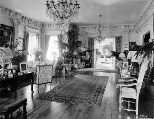Interior view of a home, probably in Denver, Colorado. Shows crystal chandeliers, oriental rugs, potted plants, lamps, furniture, drapery, decorative columns and molding.