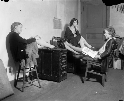 Women sit around a desk in Colorado. A woman in high heeled shoes has her feet up, by a telephone and typewriter.