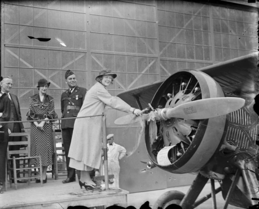 A woman christens an airplane by breaking a bottle over its propeller as men in uniform and a woman look on in possibly Denver, Colorado.