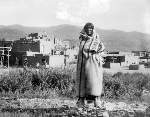 A Native American man (Taos), poses outdoors at Taos Pueblo, New Mexico. The man wears moccasins, fringed leggings, and is wrapped in a woven blanket. Adobe cluster homes and drying racks stand in the distance. The Sangre de Cristo Mountains stand in the far distance.