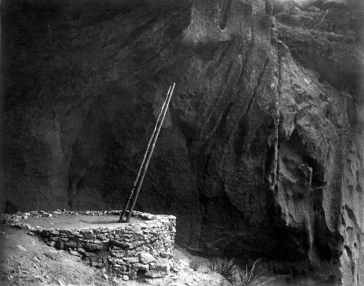 View of the entrance to a Native American (Anasazi) underground kiva in Pajarito Park (later Bandelier National Monument), New Mexico. A long wooden ladder protrudes from a square entrance to the ruins of a cylindrical stone kiva set inside a Bandelier tuff cave.  The kiva possibly stands in the side of a cliff.