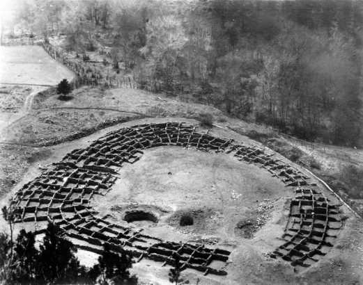 View of the Native American (Anasazi) Tyuonyi ruins in Pajarito Park (later Bandelier National Monument), New Mexico. A circular group of foundations of Pueblo style buildings circumscribe possibly three round kiva holes. A wooden rail fence separates the excavated ruins from trees and bushes growing on the adjacent mountains. A dirt road leads to the ruins.