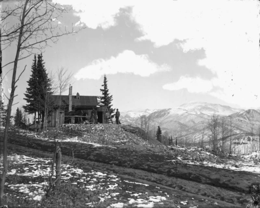 View of the Climax Mine works near Midway, Teller County, Colorado. Three men sit and stand near a wooden building with a metal roof through which a hoist protrudes. A horse stands near the building. A pile of mine tailings stands near the works. Patches of light snow cover the ground. Snow covered Pike's Peak stands in the far distance.