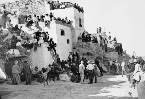 View of spectators awaiting the beginning of the Snake Dance at Walpi Pueblo, First Mesa, Arizona (Hopi Native American). Groups of men, women and children sit and stand on pueblo buildings with chimney pots as well as on adobe steps and near the dance area. Photographers and their cameras stand among the crowd.