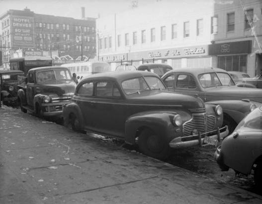 View of automobiles on Welton Street in Denver, Colorado. Background signs read: "Rock Island Lines, Route of the Rockets," "Hotel Denver, 80 Outside Rooms, $1.00 to $3.00, Thoroughly Modern, Permanent and Transient."